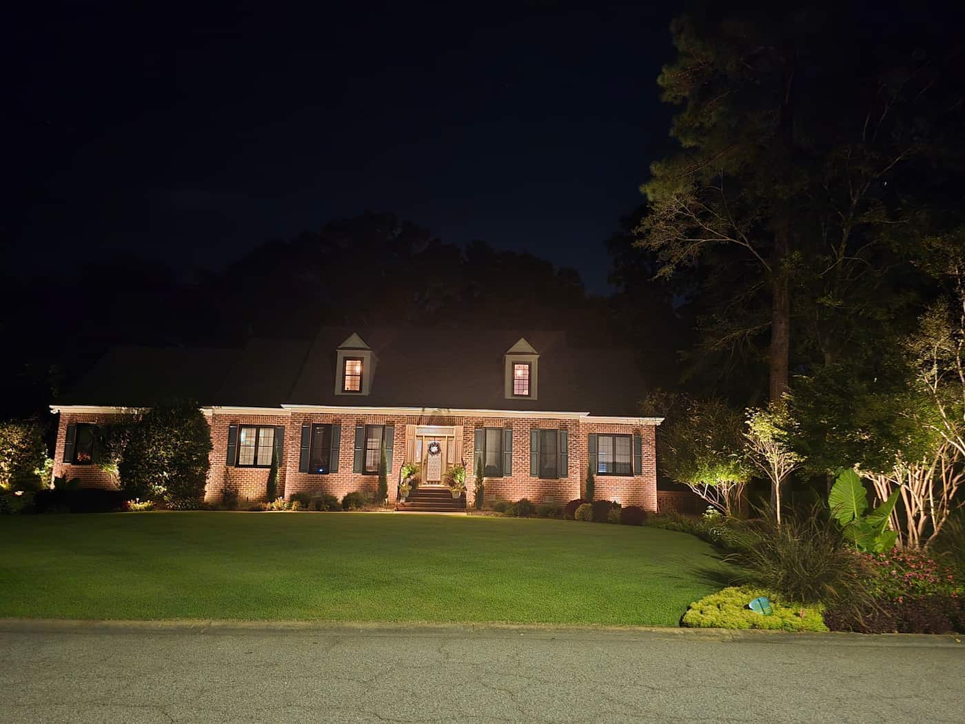 The lit facade and dormers, with trees and shrubs uplighted at the sides of the yard