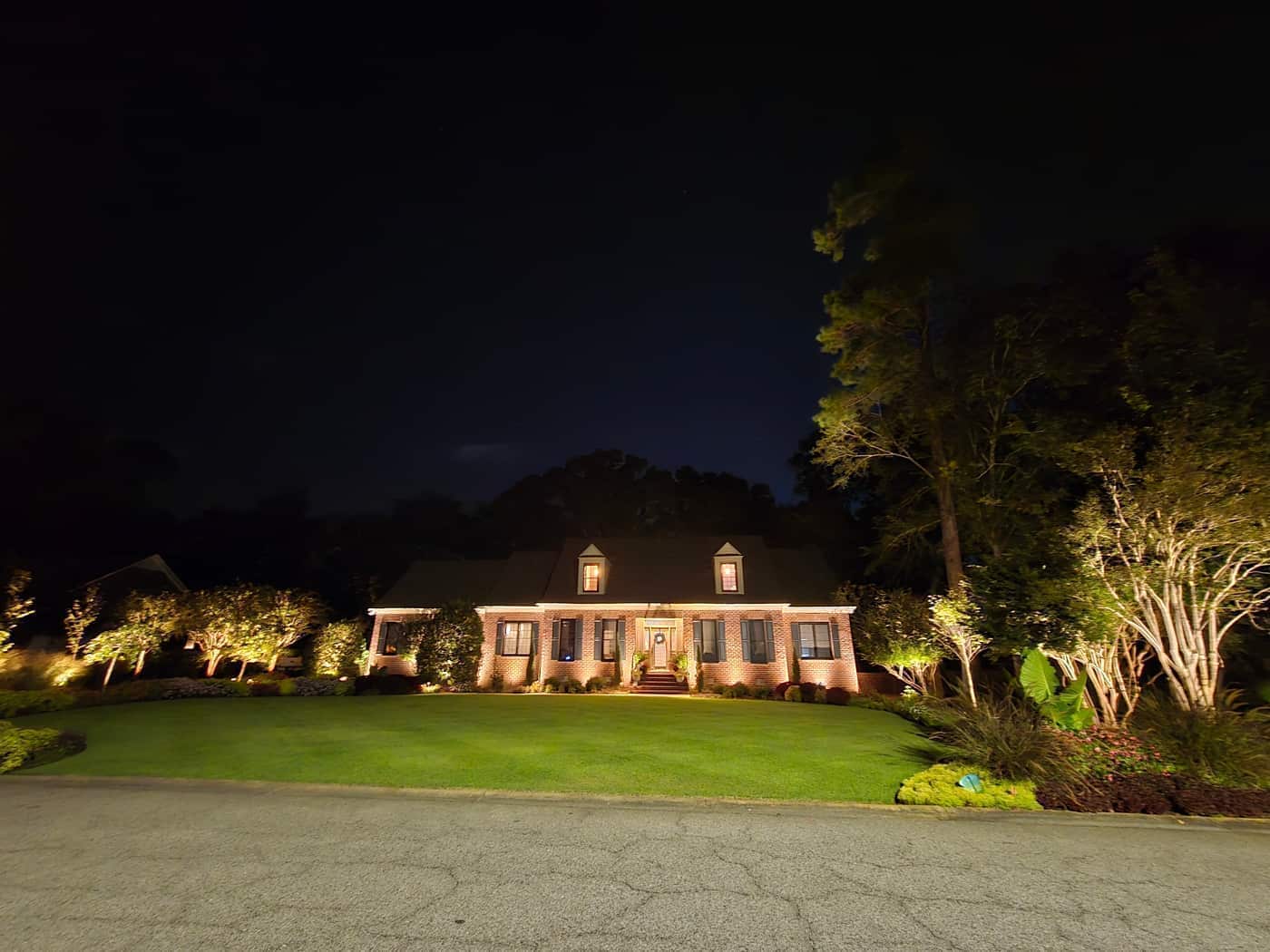 A wider night view of the brick home, lawn, and uplighted trees