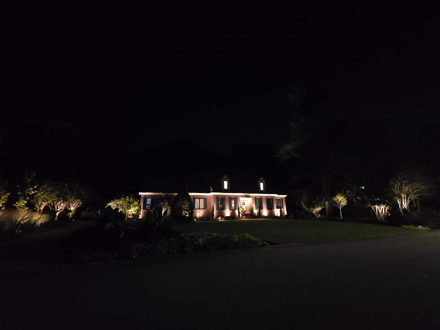 The house from the street at night, with landscape lighting on the trees along both sides of the yard