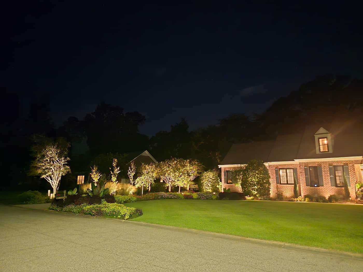 Landscape lighting on the side garden, trees, and lawn, with the brick house at the edge of the frame