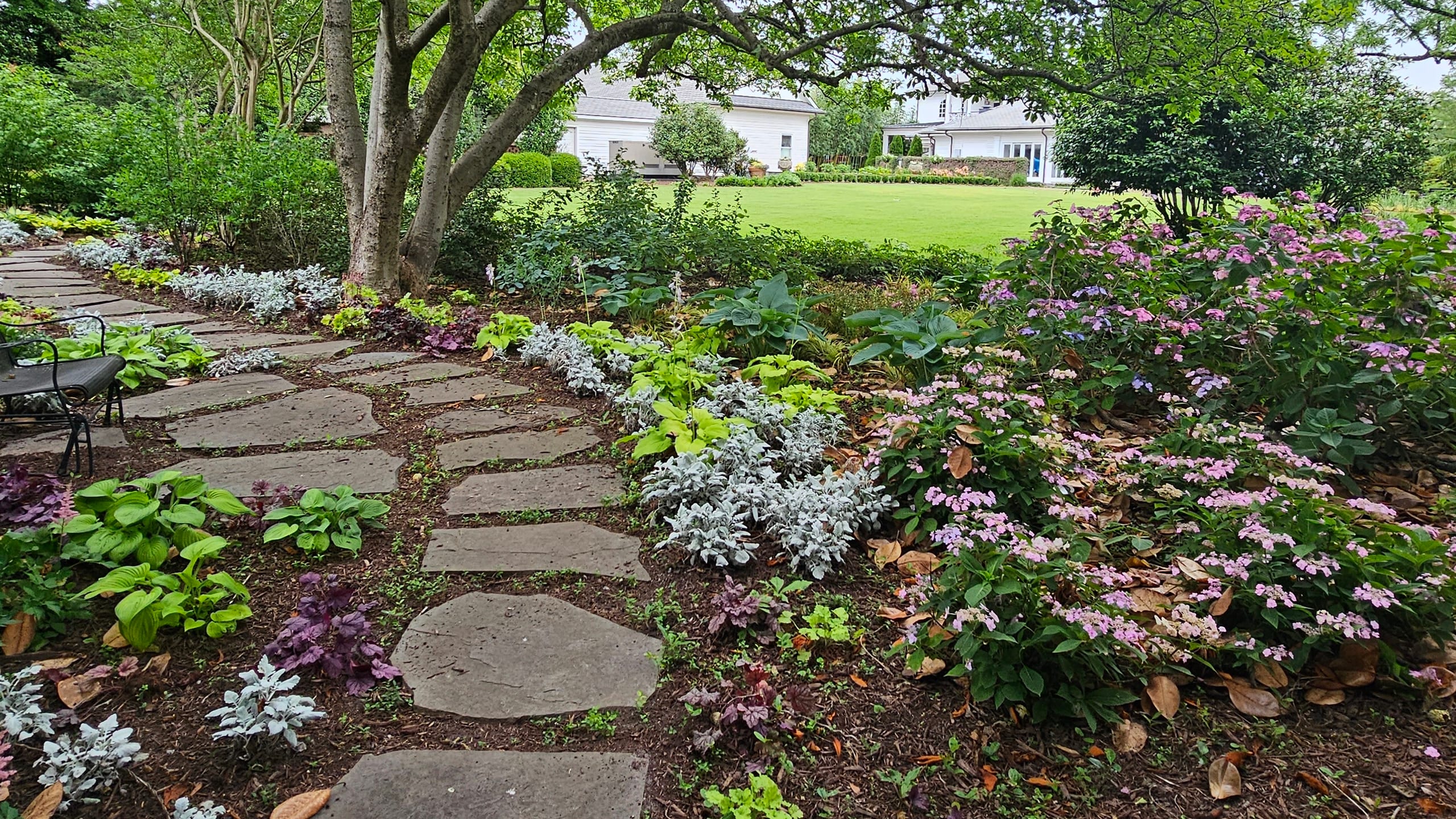 A garden path through flowering beds toward a lawn and white home